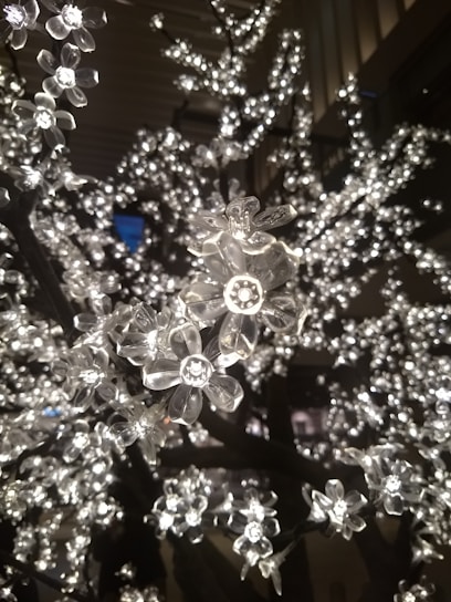 A close-up of artificial flowers made from clear plastic, illuminated by small LED lights, creating a shimmering effect. The branches appear densely covered with these glowing blossoms set against a dark background.