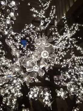 A close-up of artificial flowers made from clear plastic, illuminated by small LED lights, creating a shimmering effect. The branches appear densely covered with these glowing blossoms set against a dark background.
