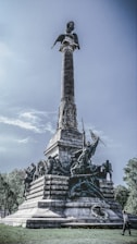 A towering monument with intricate sculptures of historical figures and symbols positioned at its base, culminating in a tall column topped by a statue. The structure is set in an open area with trees in the background, and a person is walking nearby, which emphasizes the scale of the monument.