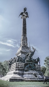 A towering monument with intricate sculptures of historical figures and symbols positioned at its base, culminating in a tall column topped by a statue. The structure is set in an open area with trees in the background, and a person is walking nearby, which emphasizes the scale of the monument.