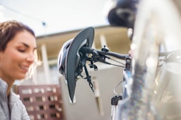 A bicycle helmet hangs on the handlebars of a bicycle. In the background, a smiling person can be seen blurred.