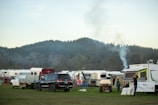 A scenic camping area with several parked caravans and SUVs is set against a backdrop of lush, forested hills. Smoke is rising from one of the parked campers, suggesting a fire or cooking inside. People are seen engaging in activities around the caravans. The atmosphere appears peaceful and organized.