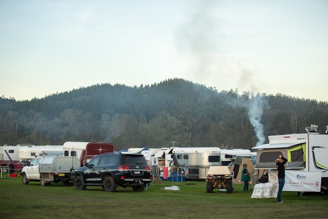 A scenic camping area with several parked caravans and SUVs is set against a backdrop of lush, forested hills. Smoke is rising from one of the parked campers, suggesting a fire or cooking inside. People are seen engaging in activities around the caravans. The atmosphere appears peaceful and organized.