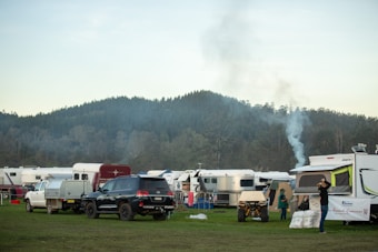 A scenic camping area with several parked caravans and SUVs is set against a backdrop of lush, forested hills. Smoke is rising from one of the parked campers, suggesting a fire or cooking inside. People are seen engaging in activities around the caravans. The atmosphere appears peaceful and organized.