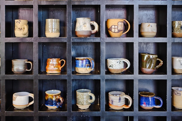 A collection of finished ceramic mugs with earthy glazes arranged on a wooden shelf, showcasing subtle variations in color.