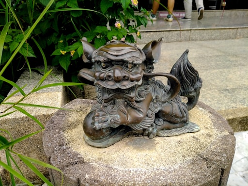 A bronze, intricately detailed statue of a mythical creature is positioned on a textured stone platform. The creature has sharp, defined features with a fierce expression, curled tail, and elaborate mane. In the background, lush green plants and flowers add vibrancy to the scene. Partially visible steps and feet suggest an outdoor setting.