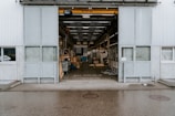 An industrial warehouse with a large open entrance showing the interior space filled with various equipment and materials. The exterior walls are made of corrugated metal, and there is a visible overhead crane inside. The building is situated on a wet pavement, indicating recent rain.