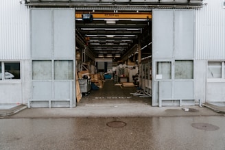 An industrial warehouse with a large open entrance showing the interior space filled with various equipment and materials. The exterior walls are made of corrugated metal, and there is a visible overhead crane inside. The building is situated on a wet pavement, indicating recent rain.