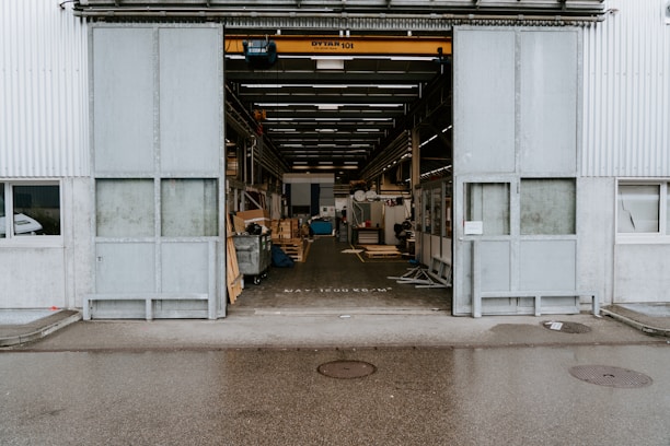 An industrial warehouse with a large open entrance showing the interior space filled with various equipment and materials. The exterior walls are made of corrugated metal, and there is a visible overhead crane inside. The building is situated on a wet pavement, indicating recent rain.