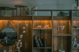 A cozy and rustic living space featuring a wooden shelf with glowing lights. It holds decorative glassware, books, and a cup. A round mirror hangs on the wooden wall, reflecting part of the room. A plant and a vase with flowers add a natural touch, while a pillow with animal prints and a cozy blanket enhance the ambiance.