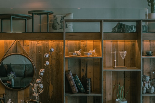 A cozy and rustic living space featuring a wooden shelf with glowing lights. It holds decorative glassware, books, and a cup. A round mirror hangs on the wooden wall, reflecting part of the room. A plant and a vase with flowers add a natural touch, while a pillow with animal prints and a cozy blanket enhance the ambiance.