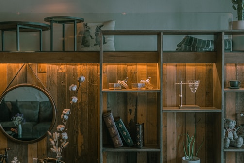 A cozy and rustic living space featuring a wooden shelf with glowing lights. It holds decorative glassware, books, and a cup. A round mirror hangs on the wooden wall, reflecting part of the room. A plant and a vase with flowers add a natural touch, while a pillow with animal prints and a cozy blanket enhance the ambiance.