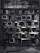 A collection of vintage televisions and electronic equipment stacked in a grid pattern against a textured stone wall. The scene is monochromatic, contributing to an industrial and retro aesthetic.