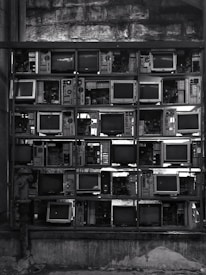 A collection of vintage televisions and electronic equipment stacked in a grid pattern against a textured stone wall. The scene is monochromatic, contributing to an industrial and retro aesthetic.