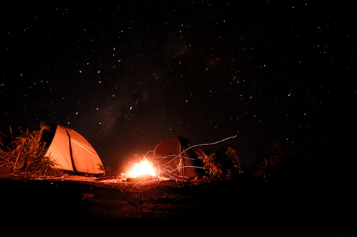 Campfire glowing under a star-filled night sky with tents nearby.