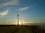 A row of wind turbines lines a coastal road at sunset, with the warm glow of the sun casting long shadows. The sky is a gradient of blue and orange, with wispy clouds scattered throughout. A wooden fence runs parallel to the road, while tall grasses sway gently in the breeze.