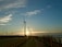 A row of wind turbines lines a coastal road at sunset, with the warm glow of the sun casting long shadows. The sky is a gradient of blue and orange, with wispy clouds scattered throughout. A wooden fence runs parallel to the road, while tall grasses sway gently in the breeze.