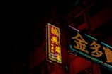 Neon signs in a city environment cast vibrant glow against a dark background, featuring intricate Chinese characters. The signs illuminate parts of the building facade with their colorful lights, creating a contrast against the night.