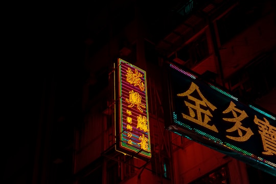 Neon signs in a city environment cast vibrant glow against a dark background, featuring intricate Chinese characters. The signs illuminate parts of the building facade with their colorful lights, creating a contrast against the night.
