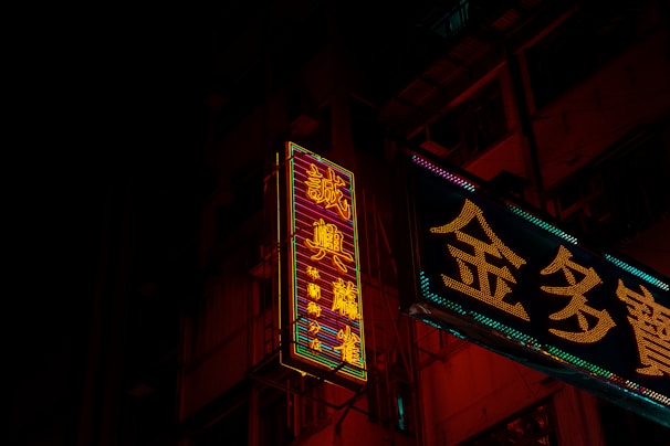 Neon signs in a city environment cast vibrant glow against a dark background, featuring intricate Chinese characters. The signs illuminate parts of the building facade with their colorful lights, creating a contrast against the night.