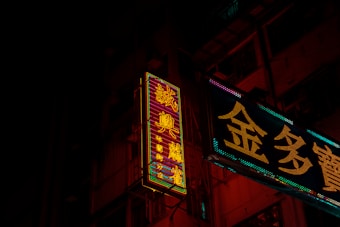 Neon signs in a city environment cast vibrant glow against a dark background, featuring intricate Chinese characters. The signs illuminate parts of the building facade with their colorful lights, creating a contrast against the night.