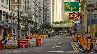 A bustling urban street with tall apartment buildings on either side, featuring air conditioning units attached to the exteriors. The street is undergoing construction, with barriers and cones marking off areas. Various signs in different languages hang above shops, indicating diverse commercial activity. A green pedestrian bridge crosses over the street further down.