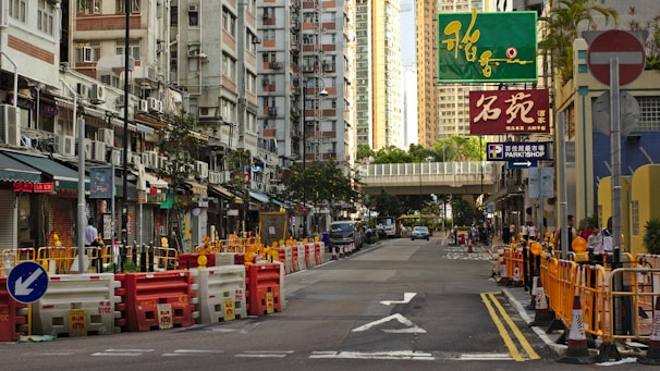 A bustling urban street with tall apartment buildings on either side, featuring air conditioning units attached to the exteriors. The street is undergoing construction, with barriers and cones marking off areas. Various signs in different languages hang above shops, indicating diverse commercial activity. A green pedestrian bridge crosses over the street further down.