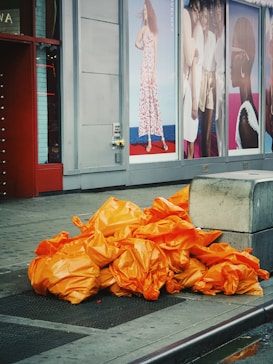 A pile of orange garbage bags is stacked on the sidewalk next to a building. Large fashion advertisements featuring women in various outfits are displayed in the background. A concrete barrier is situated nearby.