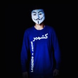 A person is wearing a white mask with a stylized smiling face and a neatly-trimmed mustache. The mask is reminiscent of the Guy Fawkes mask. The person wears a long-sleeve blue shirt with text written on it, which appears to be in English and another script. The background is black, emphasizing the figure in the foreground.