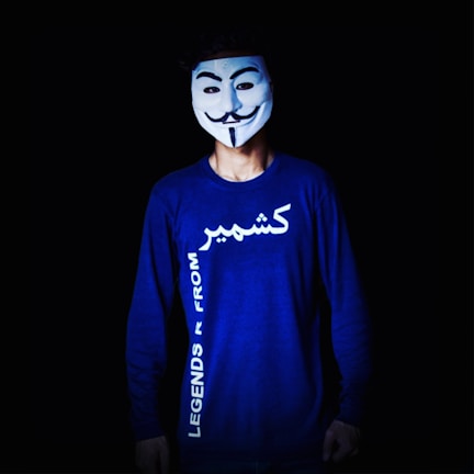 A person is wearing a white mask with a stylized smiling face and a neatly-trimmed mustache. The mask is reminiscent of the Guy Fawkes mask. The person wears a long-sleeve blue shirt with text written on it, which appears to be in English and another script. The background is black, emphasizing the figure in the foreground.