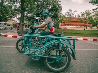A person wearing a helmet is riding a motorbike with an attached sidecar designed for carrying goods. The motorbike is traveling on a road with a red and white painted curb. In the background, there is a traditional building with a red roof and some trees, and a van is visible on the road.