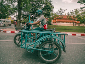A person wearing a helmet is riding a motorbike with an attached sidecar designed for carrying goods. The motorbike is traveling on a road with a red and white painted curb. In the background, there is a traditional building with a red roof and some trees, and a van is visible on the road.