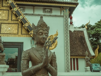 A bronze statue of a deity with intricate detailing sits in front of a richly decorated temple structure. The temple facade features ornate gilded patterns and traditional architectural elements, including a steeply pitched roof adorned with sculptures. Lush green trees can be seen in the background under a cloudy sky.