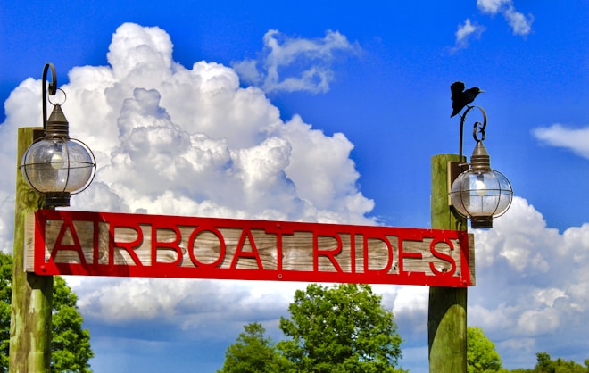 A red wooden sign with 'AIRBOAT RIDES' carved out stands between two rustic lanterns mounted on wooden posts. A bird is perched on the top of the right lantern. The background features lush green trees and a bright blue sky filled with large, fluffy white clouds.