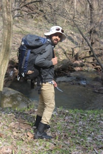 A person is standing near a small stream in a forested area, wearing a wide-brimmed hat, sunglasses, a large backpack, and holding a small tool or knife. The individual is dressed in outdoor gear, suggesting a hiking or camping trip. The ground is covered with fallen leaves and some patches of green grass, and bare tree branches are visible in the background.