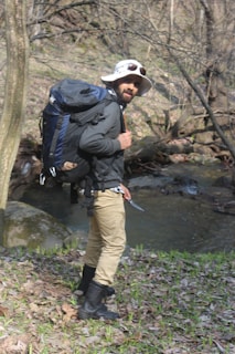 A person is standing near a small stream in a forested area, wearing a wide-brimmed hat, sunglasses, a large backpack, and holding a small tool or knife. The individual is dressed in outdoor gear, suggesting a hiking or camping trip. The ground is covered with fallen leaves and some patches of green grass, and bare tree branches are visible in the background.