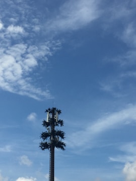 A cell tower designed to look like a tree, standing against a backdrop of blue sky with scattered clouds. The tower is adorned with artificial branches to blend in with the natural environment.