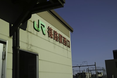 A beige concrete wall with 'JR' and Japanese characters in green and dark red. There is a metal pipe along the wall and part of a doorframe is visible. In the background, there are electrical wires and poles against a clear blue sky.