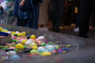 A variety of colorful plastic toys float in a clear rectangular water basin. The toys include ball-shaped objects and animals in bright colors such as yellow, blue, pink, and green. There is a partial view of a person standing nearby, wearing a blue striped garment.