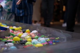 A variety of colorful plastic toys float in a clear rectangular water basin. The toys include ball-shaped objects and animals in bright colors such as yellow, blue, pink, and green. There is a partial view of a person standing nearby, wearing a blue striped garment.