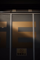 A large window is partially covered by a metallic frame and wall, casting a shadow over it. The letters 'SE' in bold are visible on the upper portion with a dark background, while a small sign featuring a bicycle with a prohibition symbol is located at the bottom.