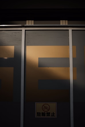 A large window is partially covered by a metallic frame and wall, casting a shadow over it. The letters 'SE' in bold are visible on the upper portion with a dark background, while a small sign featuring a bicycle with a prohibition symbol is located at the bottom.