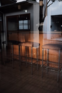 A reflective view of a modern interior setting, featuring a row of four tall wooden bar stools with dark cushions. The stools are positioned along a wooden counter. There's a large window reflecting parts of the surroundings, including a tree and some hanging lamps. The ambient lighting is soft, creating a cozy atmosphere.
