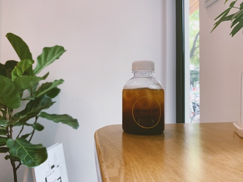 A bottle filled with a dark iced beverage sits on a wooden table. The label reads 'sensory cells.' A lush green indoor plant is visible in the background next to a window revealing a bit of the outside. Soft natural light filters through, creating a calm atmosphere.