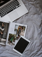A laptop with an open keyboard lies on a rumpled, light-colored fabric surface. Beside it is an open fashion magazine displaying images of people in stylish clothing. Nearby, a tablet with a black screen rests flat on the fabric.