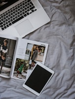 A laptop with an open keyboard lies on a rumpled, light-colored fabric surface. Beside it is an open fashion magazine displaying images of people in stylish clothing. Nearby, a tablet with a black screen rests flat on the fabric.