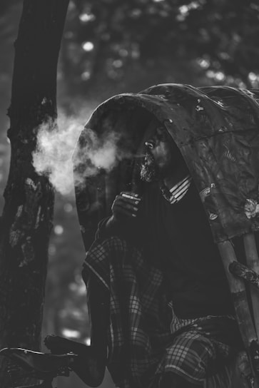 A person is seated in a rickshaw, exhaling smoke. The setting suggests a serene yet contemplative mood, with the individual partially covered by the rickshaw's canopy. The image is rendered in black and white, highlighting the texture and contrasts of the scene.
