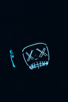 A dark, eerie setting with a glowing, stylized face mask that has Xs for eyes and a stitched mouth design, outlined in neon blue.