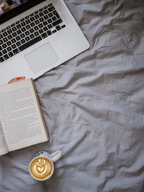 A cozy arrangement featuring an open book, a laptop keyboard, and a cup of cappuccino with latte art on a textured gray bedspread. The composition suggests a comfortable setting for reading or working from home.