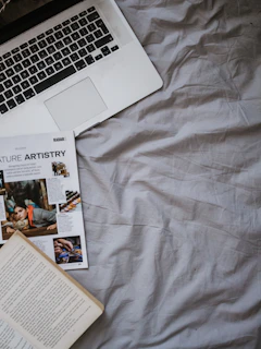A laptop partially visible in the upper left corner rests on a slightly wrinkled gray bedsheet. Below the laptop, an open magazine showcases various articles and images. An open book with text fills part of the lower left corner.